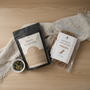 Vue de dessus d'un pack bien-être composé d'un sachet de "Tisane Maternitea" noir et d'un sachet transparent de "Racines de Khamaré", disposés sur une table en bois clair avec un linge en coton beige et un petit bol de plantes séchées.
